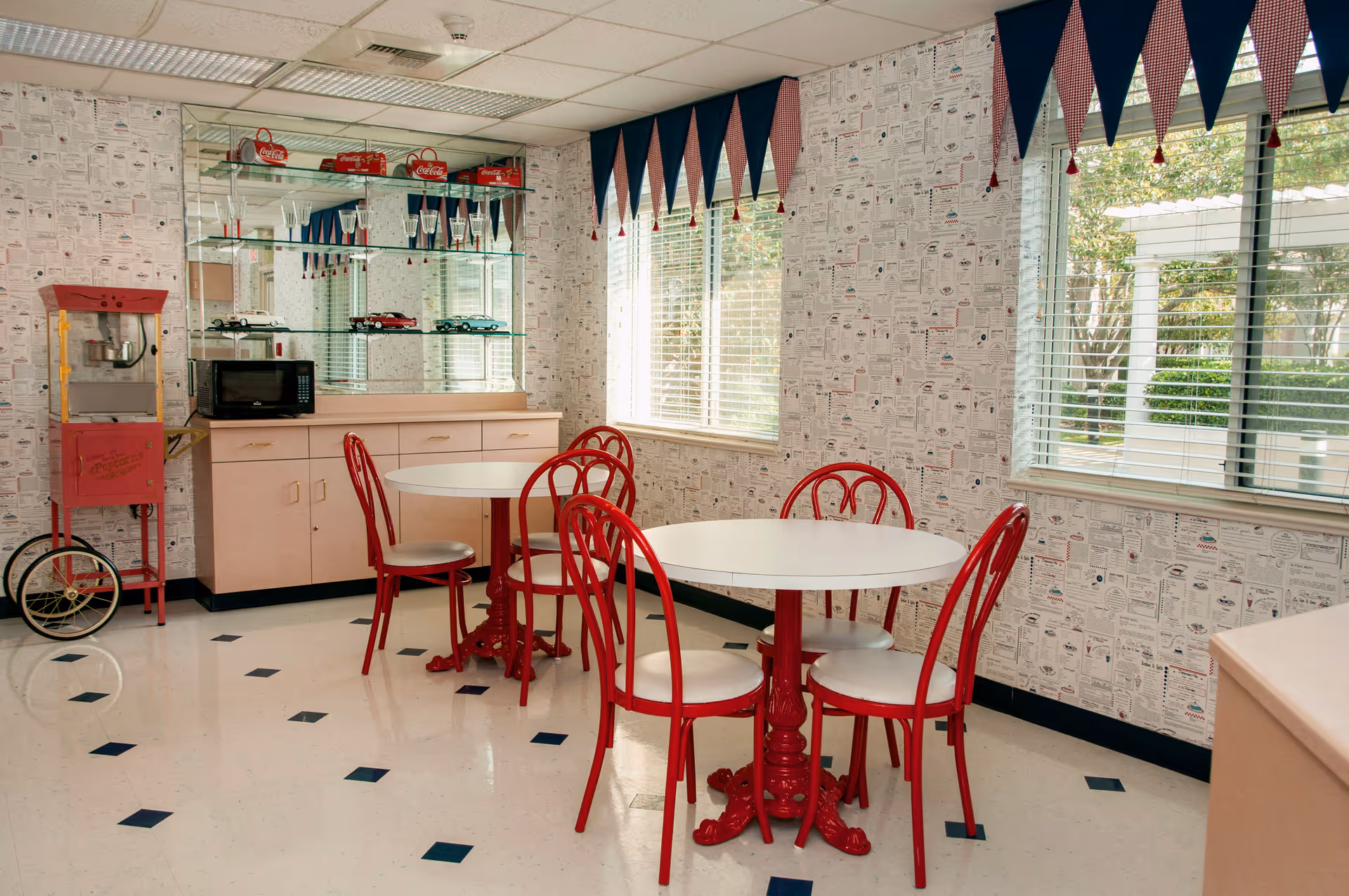 Bright retro-style dining area with round white tables, red chairs, a popcorn cart and display shelves by windows with blue pennant valances.