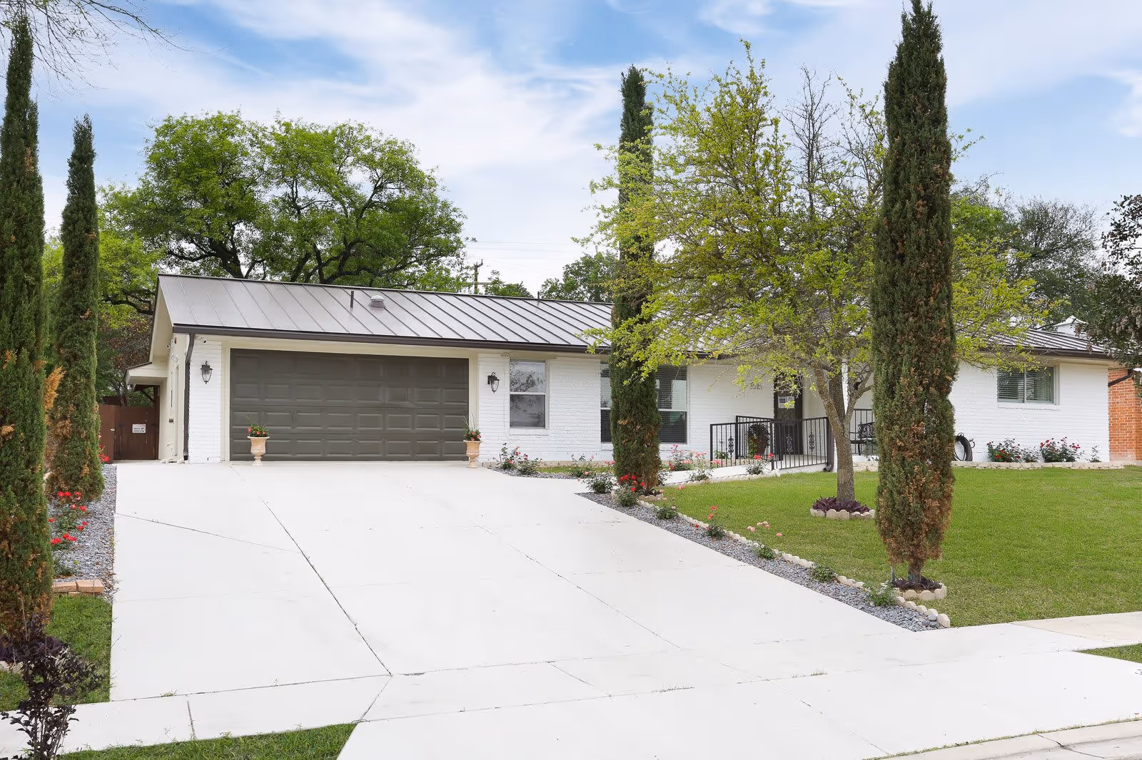 Front exterior view of a single-story white brick house with a dark gray metal roof and a large driveway leading to a double garage door. The yard features green grass, several tall narrow trees, a flowering tree, and flower beds with red and pink flowers.