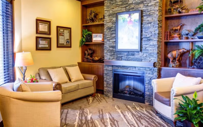 Cozy living room area with a stone fireplace in the center, flanked by built-in wooden shelves displaying decorative items and plants. There is a beige loveseat with two cushions, a matching armchair, a floor lamp, and a patterned carpet. The room has warm lighting and a window with blinds on the left side.