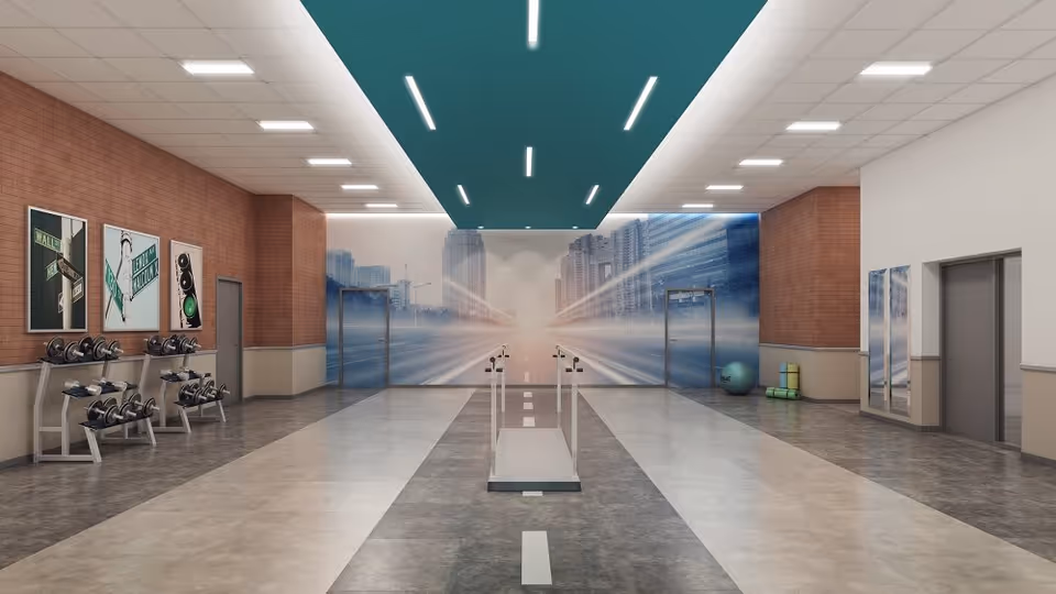 A spacious indoor exercise room with a modern design. The room features a set of dumbbells on racks along the left wall, a treadmill in the center, and exercise balls and mats in the back right corner. The walls have a mix of brick and plain finishes, with large cityscape murals on the far wall. The ceiling has recessed lighting with a teal accent panel running down the middle.