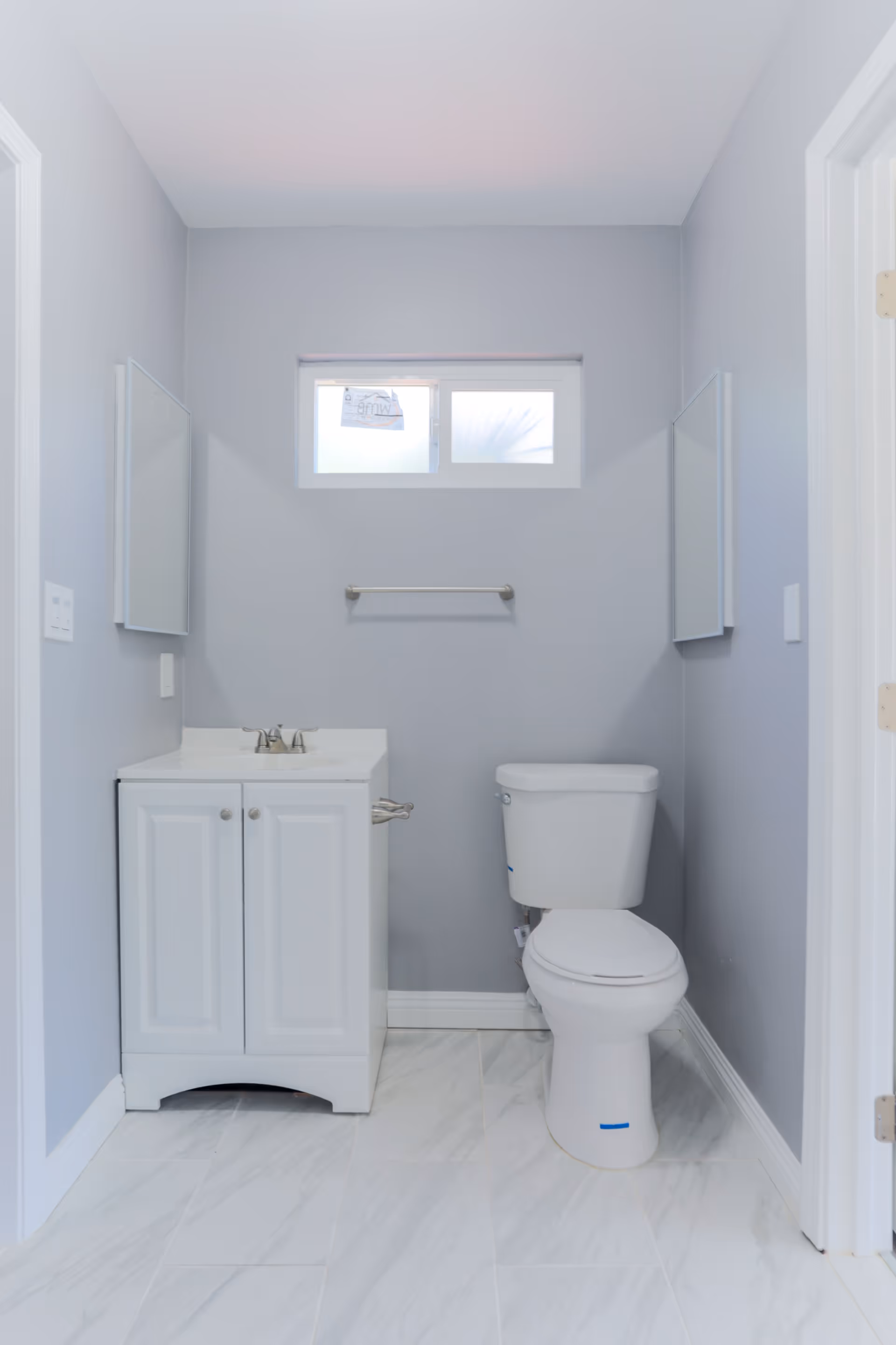 A small bathroom with light gray walls and white marble tile flooring. There is a white vanity with a sink and two mirrors on either side above it. Next to the vanity is a white toilet. A small frosted window is above the toilet with a towel bar mounted on the wall beneath it.