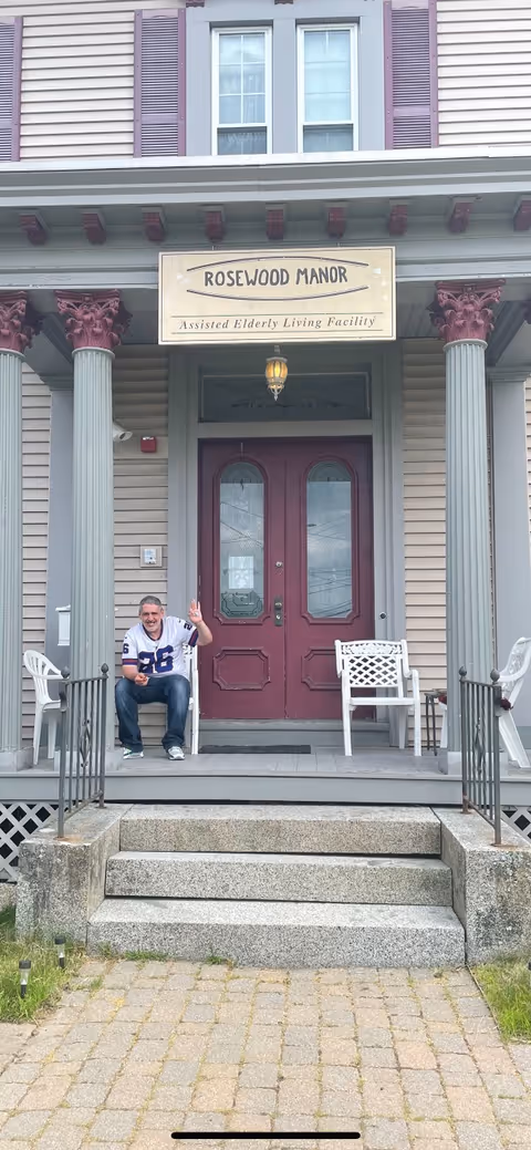 Front entrance of Rosewood Manor Assisted Elderly Living Facility showing a man sitting on the porch steps waving. The building has two large columns, a maroon double door, and a sign above the door with the facility name. There are white chairs on the porch and stone steps leading up to it.