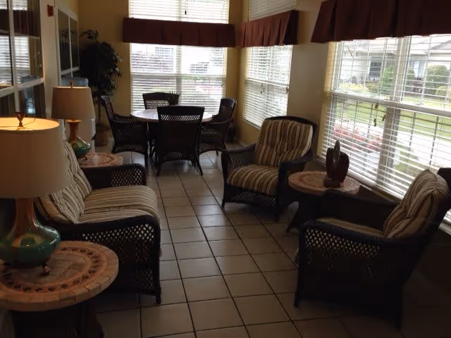 A sunlit sitting area with wicker furniture including four cushioned armchairs and a round table with a lamp. In the background, there is a dining table with four chairs near large windows with blinds, allowing natural light to fill the room. The floor is tiled and there are brown valances above the windows.