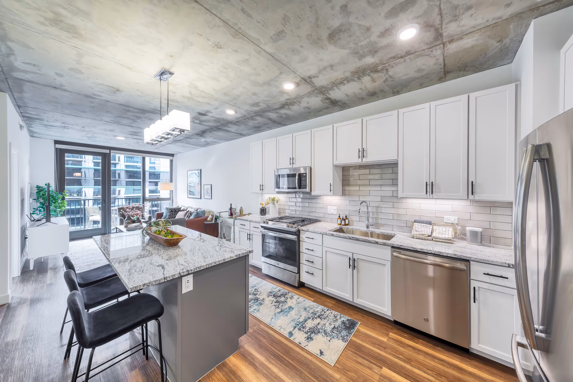 Modern kitchen and living area in an apartment with white cabinets, stainless steel appliances, a marble countertop island with three black bar stools, wooden flooring, and large windows leading to a balcony.