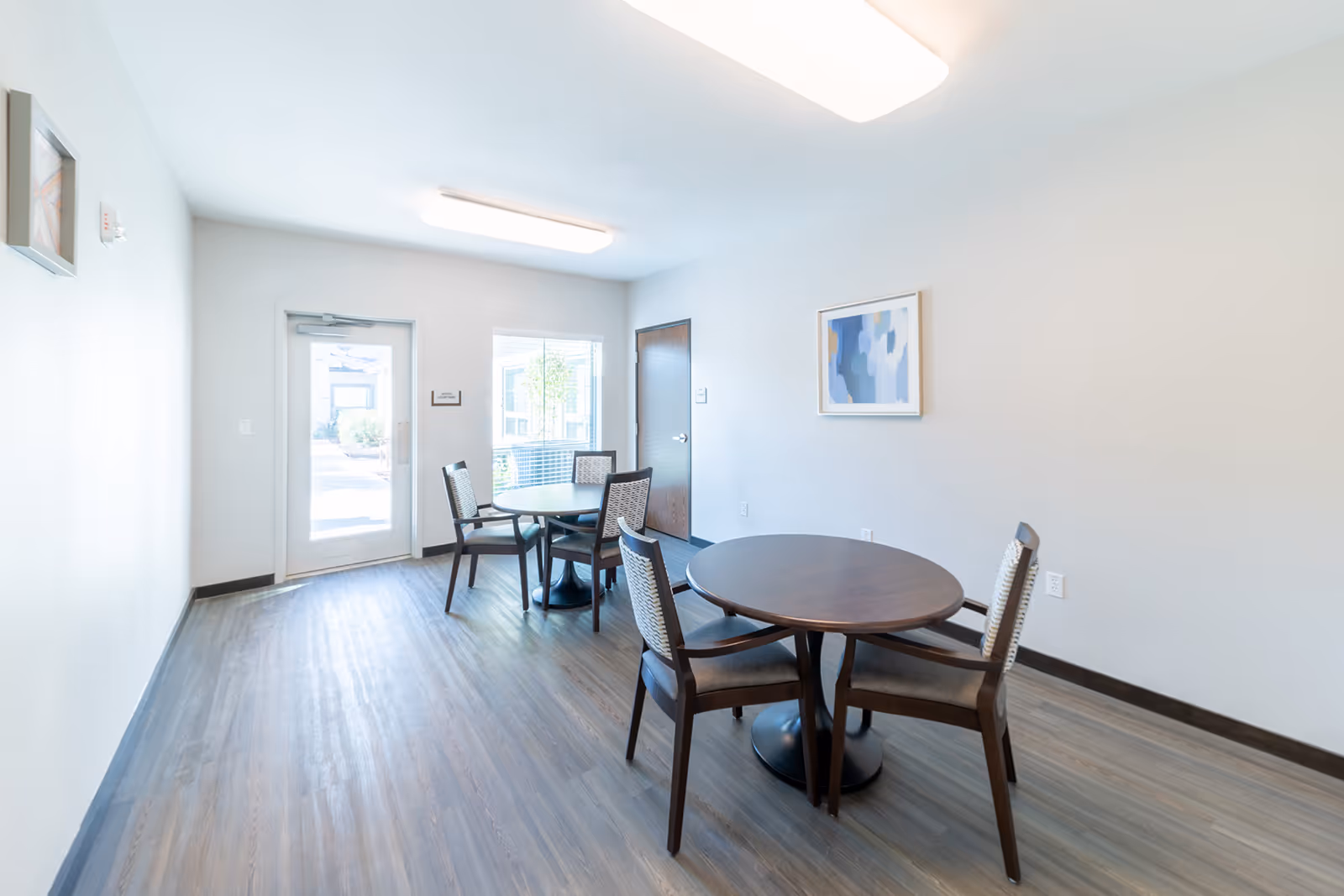 Bright common room with two round wooden tables and several chairs, light wood flooring, and a door and window letting in natural light.