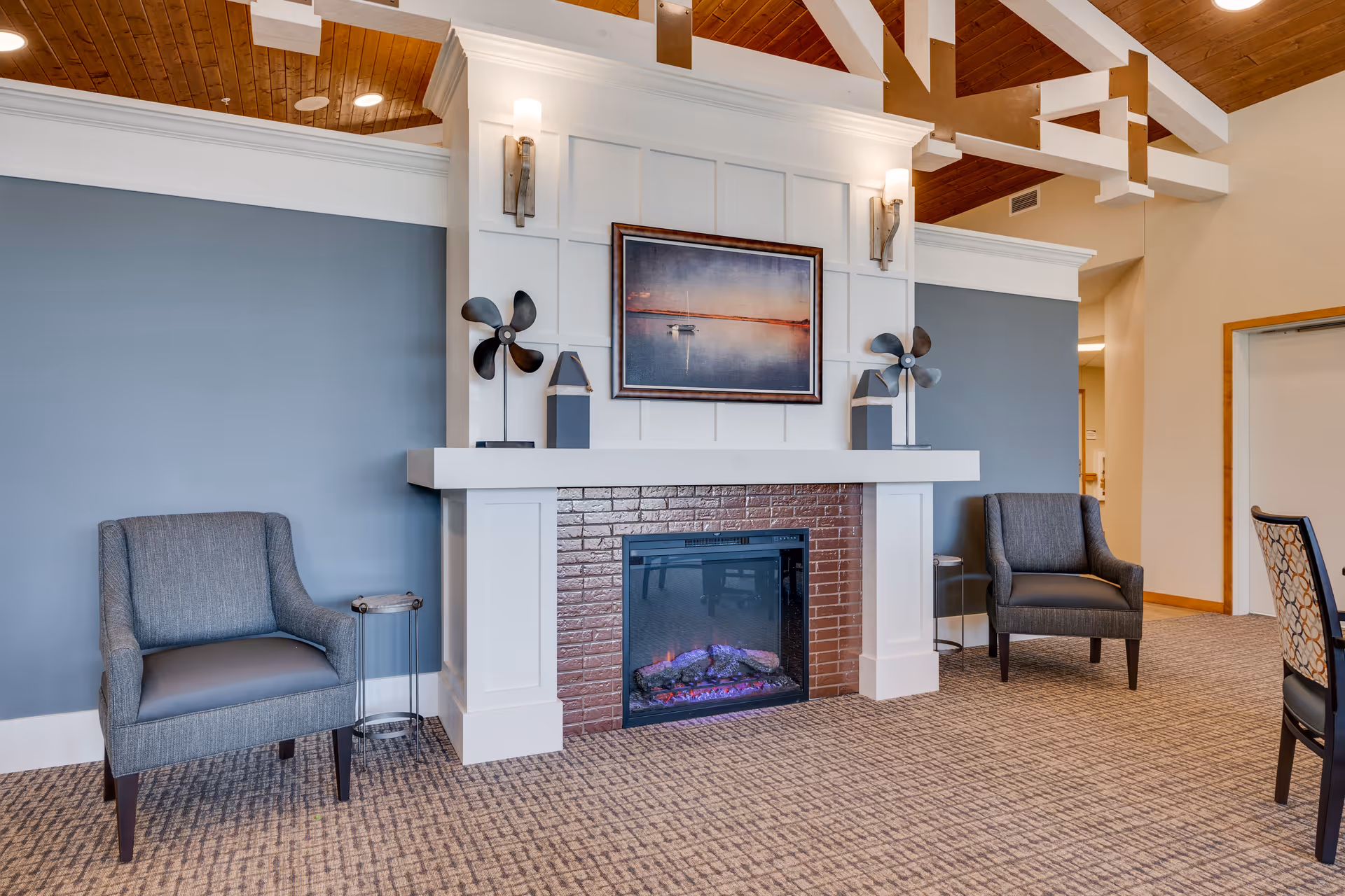 A cozy senior living common area featuring a white fireplace with a brick interior and an electric fire. Above the fireplace is a framed picture of a boat on calm water at sunset. On either side of the fireplace are two gray armchairs with small side tables next to them. The walls are painted blue and white, and the ceiling has exposed wooden beams with a wood finish.