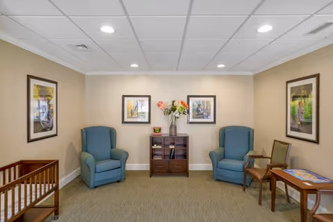 A small sitting area in a senior living facility with two blue upholstered armchairs, a wooden side table with a puzzle on it, a wooden cabinet with flowers in a vase on top, and framed artwork on the beige walls.