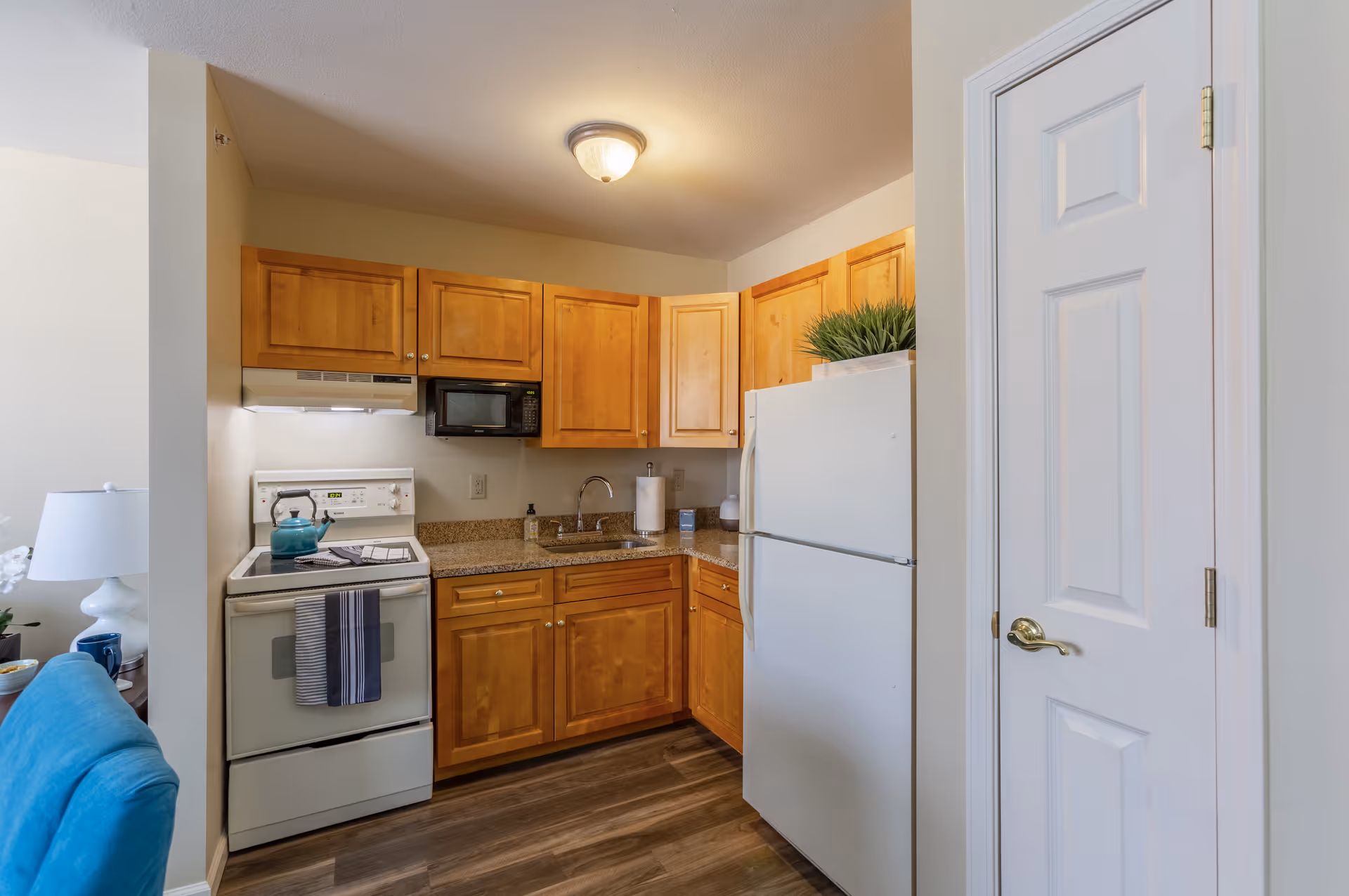A compact kitchen with wooden cabinets, a white refrigerator, a white stove with a teal kettle on it, a microwave above the stove, a sink with a paper towel holder, and a small plant on top of the refrigerator. The kitchen has a ceiling light fixture and wood-style flooring. A white door with a gold handle is visible on the right side.