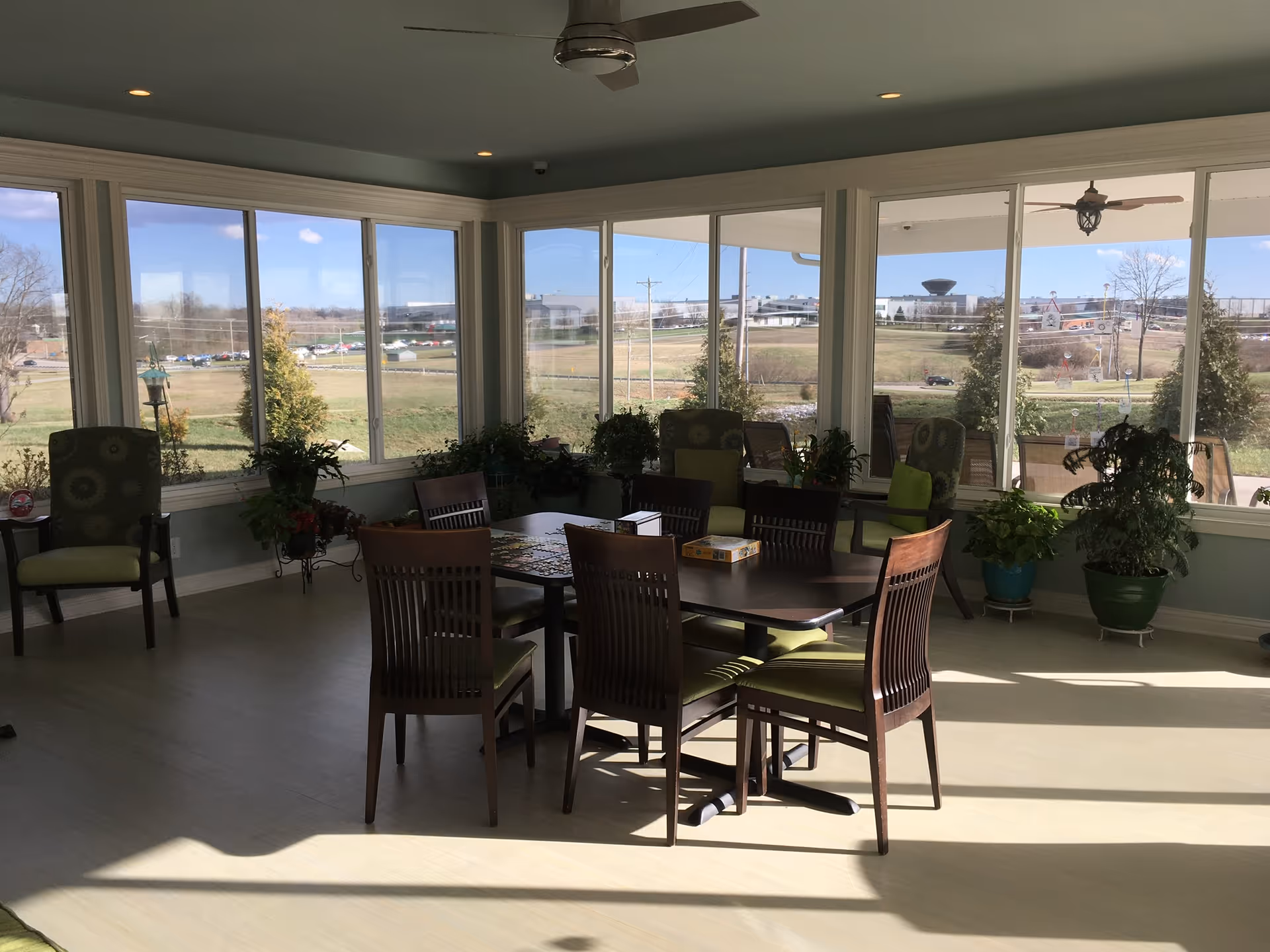 A bright sunroom with large windows showing an outdoor view. Inside, there is a dark wooden table with six chairs around it, some with green cushions. Several potted plants are placed near the windows and around the room. The ceiling has a fan and recessed lighting.