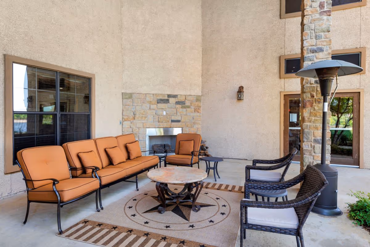 Outdoor covered patio area with cushioned seating including a sofa and chairs arranged around a round coffee table on a decorative rug. There is a stone fireplace on the wall and a tall outdoor heater near a stone pillar. Large windows and glass doors are visible in the background.