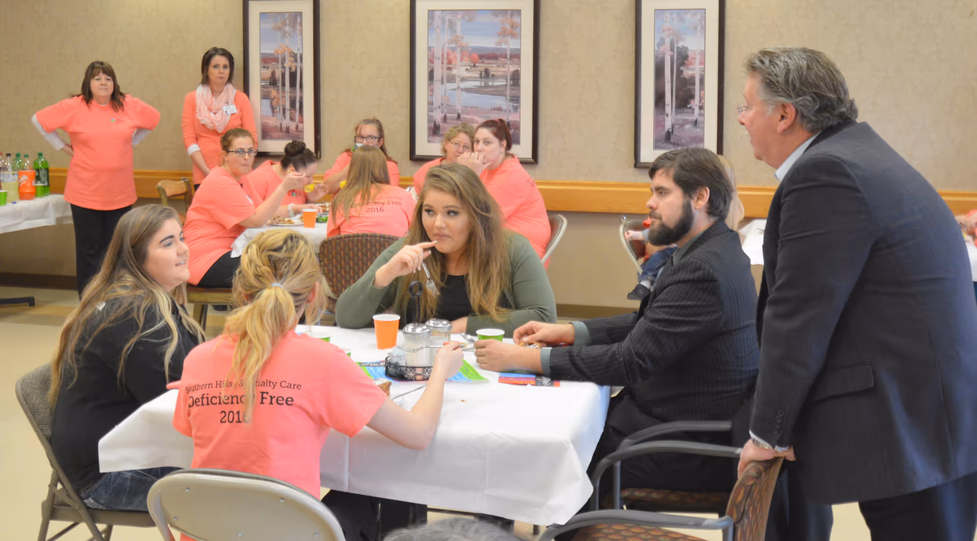 People sit and stand around a table in a communal dining area, with several staff wearing matching pink shirts.