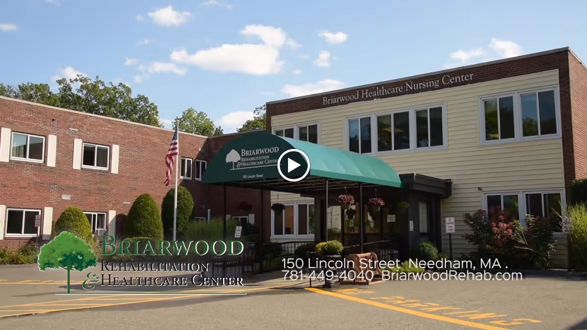 Exterior view of Briarwood Rehabilitation & Healthcare Center, a two-story building with a brick and beige facade, a green awning over the entrance, and an American flag on a flagpole in front. The sky is blue with some clouds.