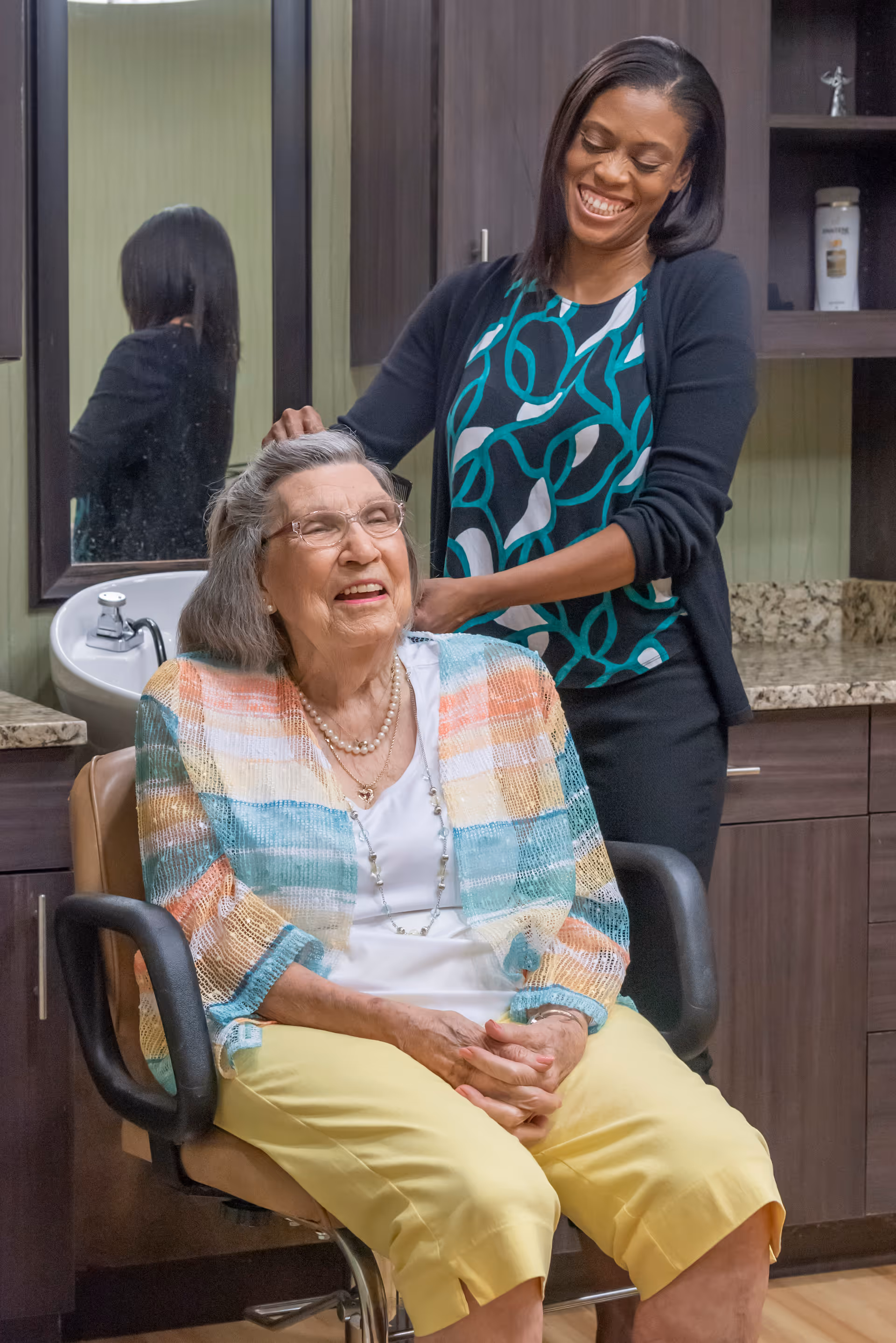 An elderly woman with glasses and a colorful cardigan sits in a salon chair smiling while a younger woman standing behind her styles her hair in a room with dark wood cabinets and a granite countertop.