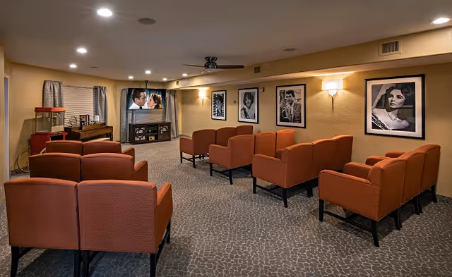 A cozy media room with rows of orange armchairs facing a wall-mounted TV and framed black-and-white portraits on the walls.