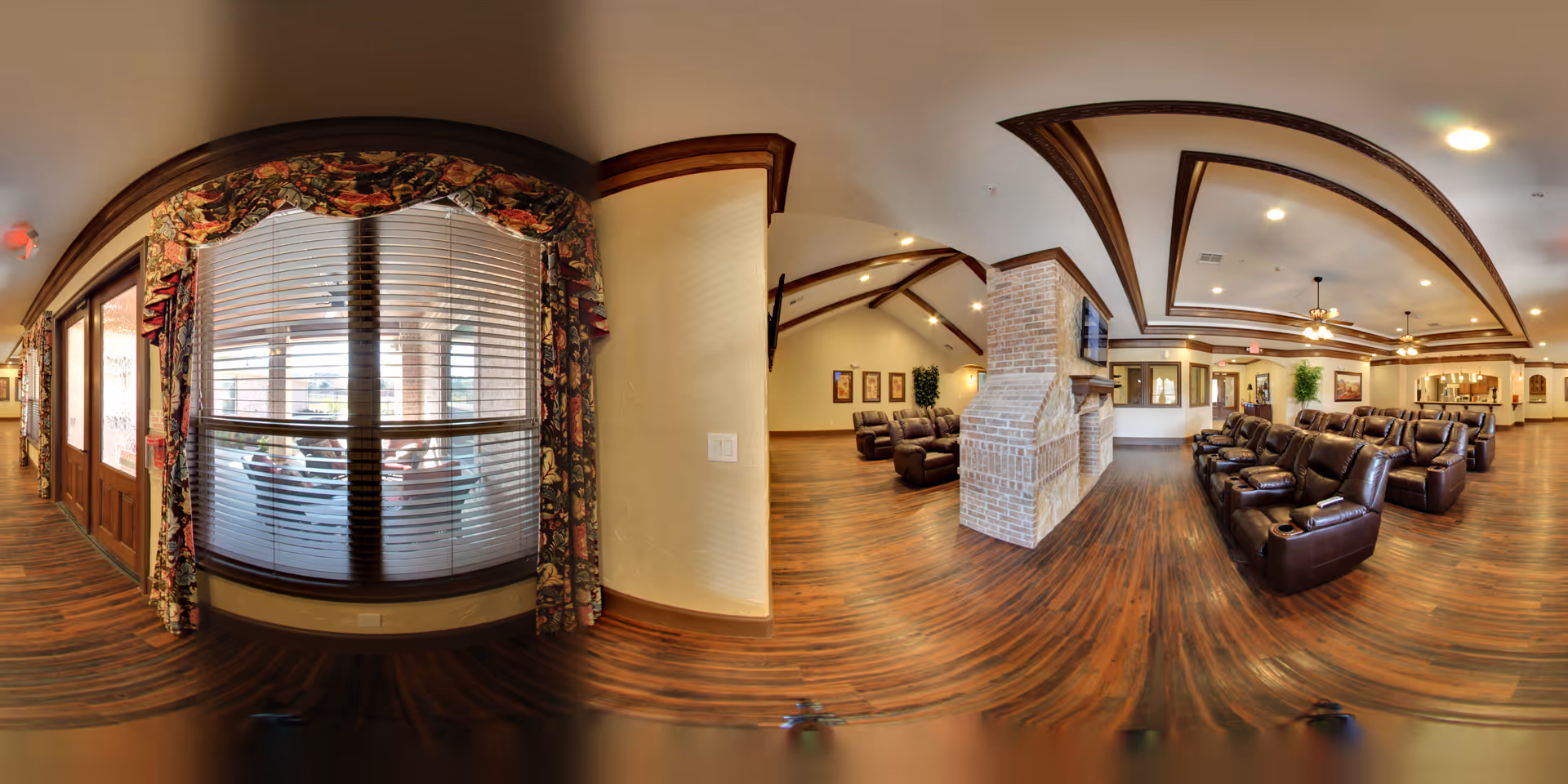 Panoramic common lounge with leather recliners, wood floors, a central brick fireplace, and large windows with blinds and floral valances.