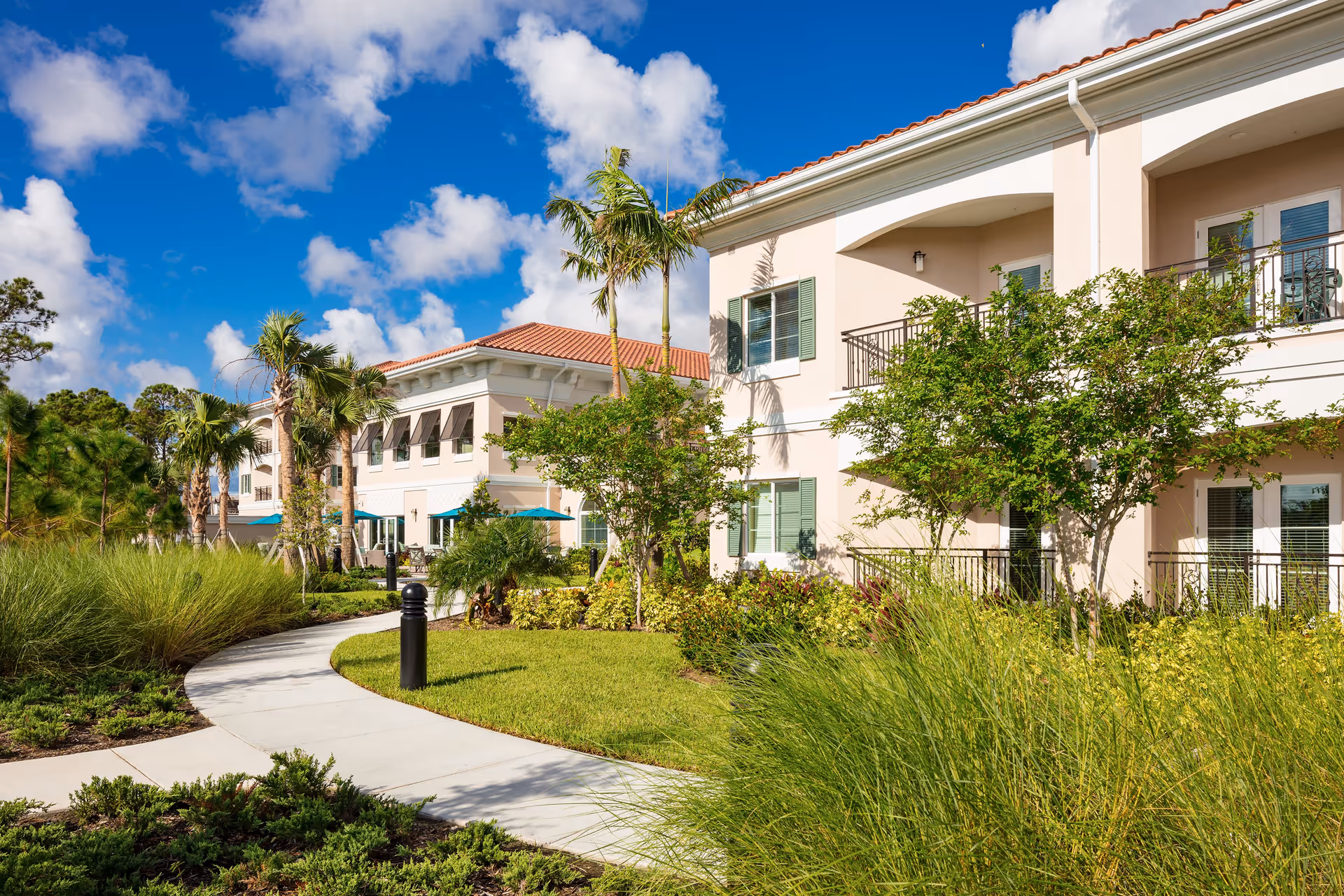 Exterior view of HarborChase of Palm Beach Gardens senior living facility with a paved walkway, lush green landscaping, palm trees, and a two-story building under a bright blue sky with scattered clouds.