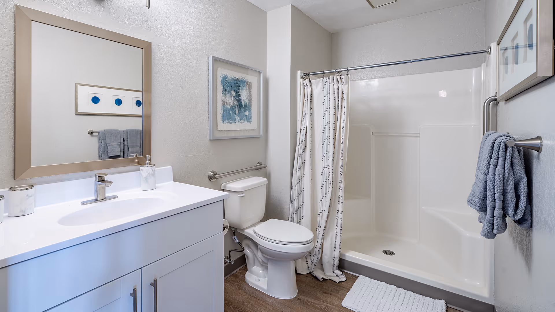 A clean and modern bathroom featuring a white vanity with a sink and faucet, a large mirror above the sink, a toilet, and a walk-in shower with a white curtain. The walls are light-colored with framed artwork, and there are gray towels hanging on towel bars. The floor has a wood-like finish.