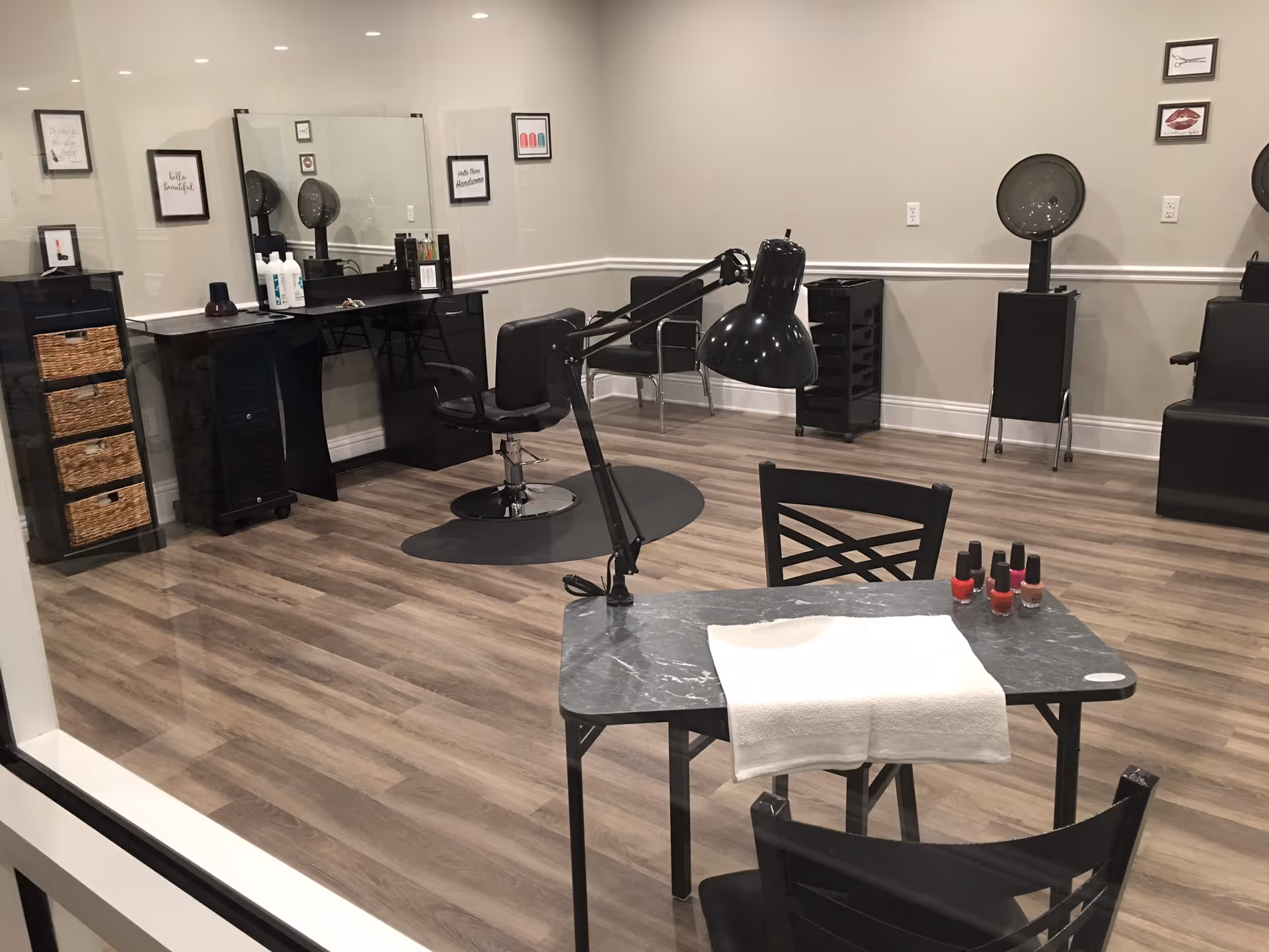 Interior view of a salon area in an assisted living facility with a manicure table in the foreground holding nail polish bottles and a towel, salon chairs, hair dryers, mirrors, and framed wall art on light-colored walls with wood flooring.