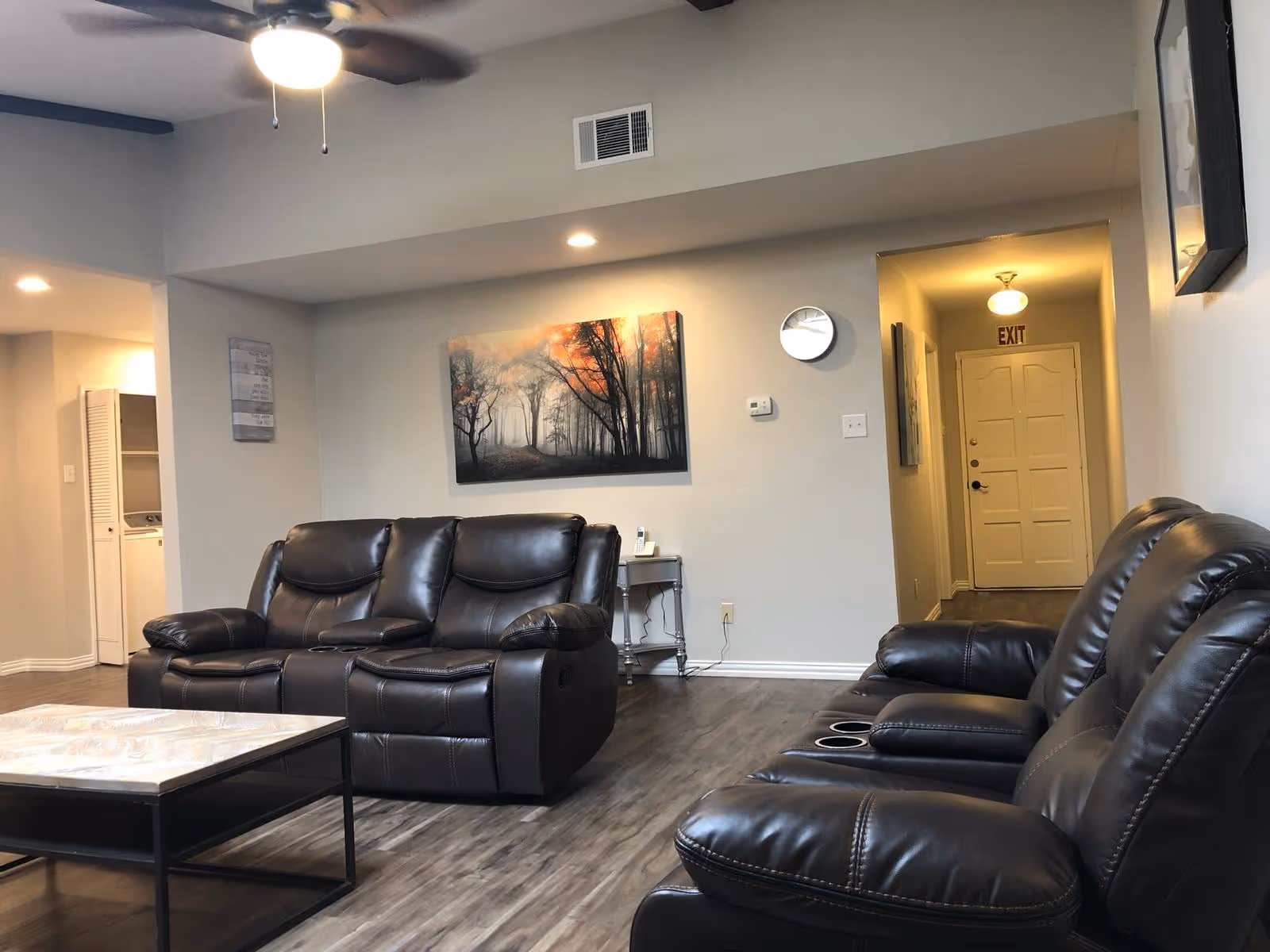 Cozy senior living lounge with black leather seating, coffee table, wall art, and hallway.