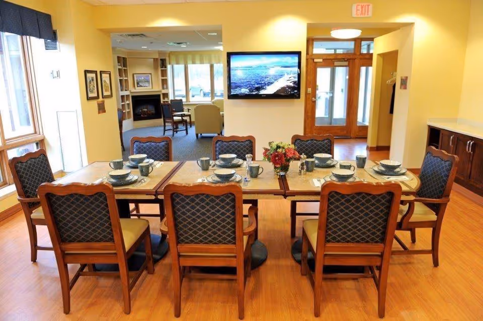 A dining area in a senior living facility with a rectangular table set for eight people. The table has matching chairs with cushioned seats and backs, and is set with plates, bowls, cups, and silverware. A small flower arrangement is in the center of the table. The room has wooden flooring, yellow walls, and large windows letting in natural light. In the background, there is a wall-mounted TV, a fireplace, and a seating area with armchairs.