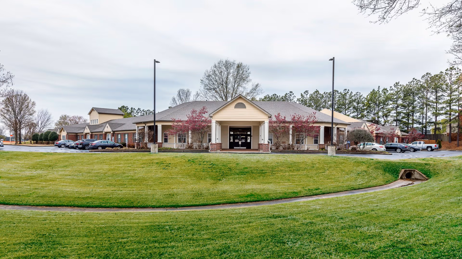 Exterior view of Hearthstone Assisted Living facility showing a single-story building with a covered entrance, surrounded by a well-maintained lawn and several parked cars. Trees and lampposts are visible around the building under a cloudy sky.