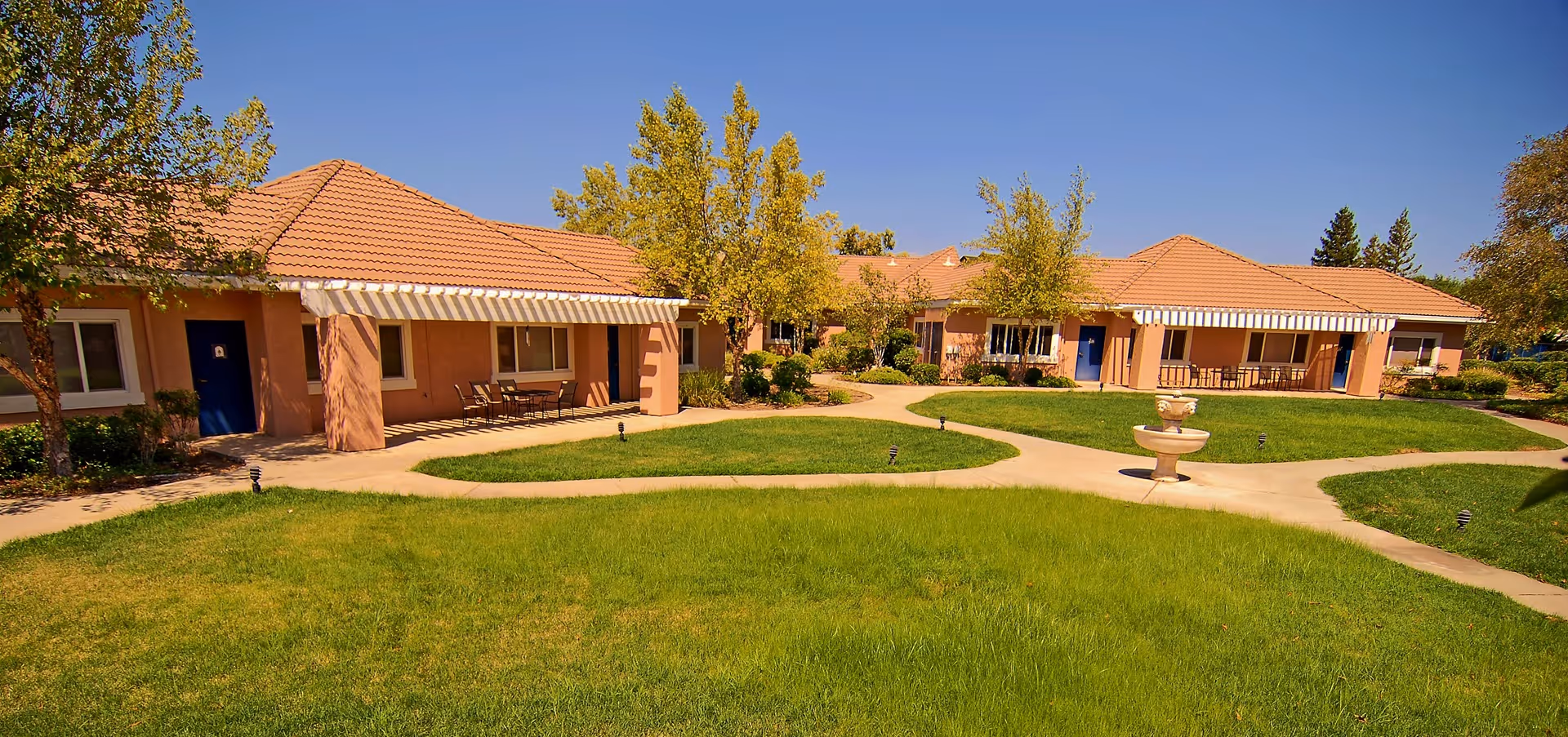 A sunny outdoor courtyard area at Roseleaf Gardens featuring a green lawn, paved walkways, several small trees, and a central water fountain. The surrounding buildings have terracotta tiled roofs and beige walls with blue doors and striped awnings over patio seating areas.