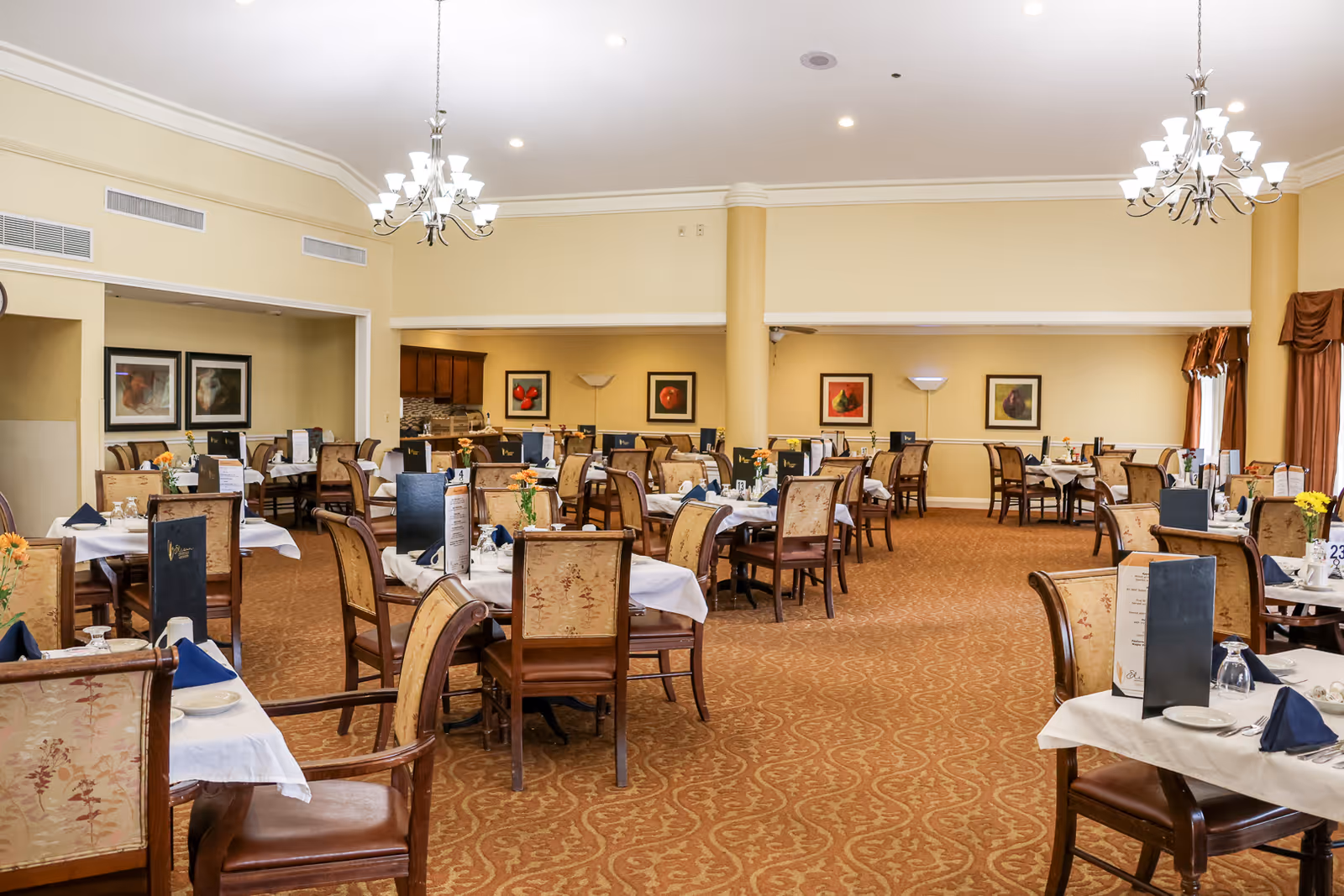 A spacious dining room with multiple tables covered in white tablecloths, each set with plates, glasses, napkins, and menus. The room features beige walls, carpeted floors with a patterned design, chandeliers hanging from the ceiling, and framed artwork on the walls. Chairs with cushioned seats and backs surround the tables, and there are windows with brown curtains on the right side.