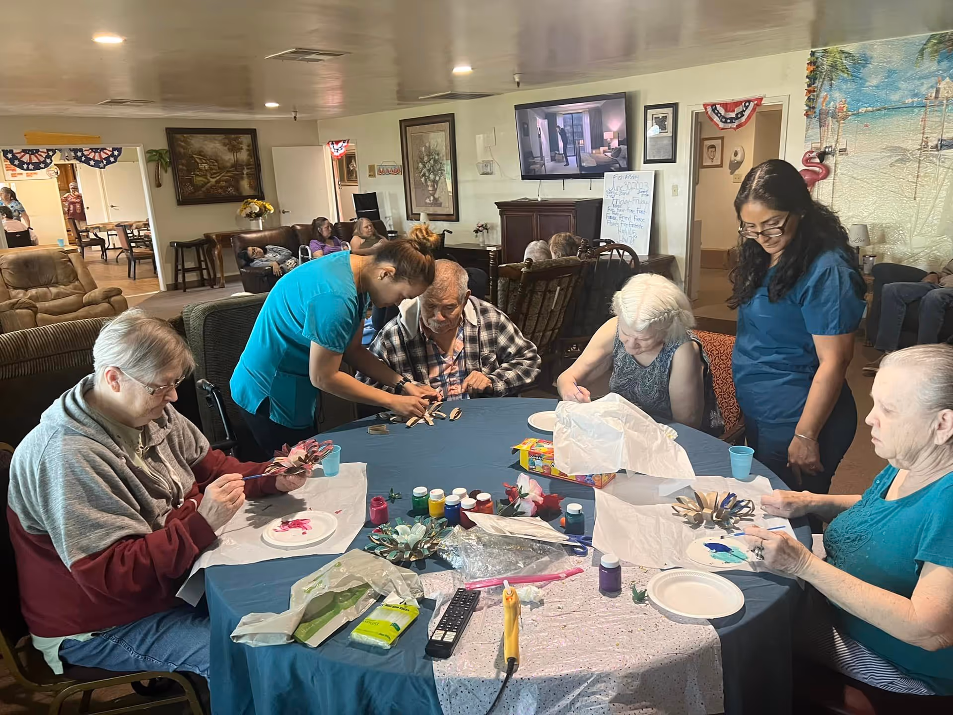 A group of elderly individuals seated around a table engaged in a painting and craft activity, assisted by two caregivers in a cozy common room decorated with paintings and patriotic bunting. Various paint bottles, brushes, and craft materials are spread on the table.