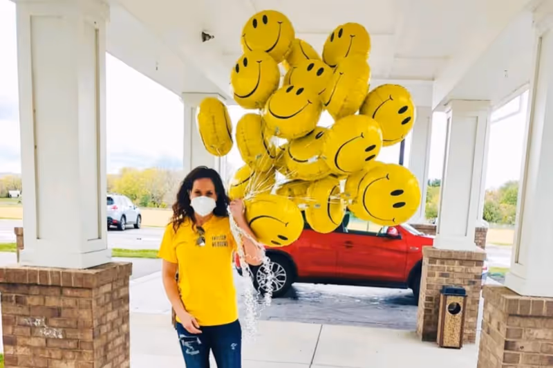 A woman wearing a yellow English Meadows t-shirt and a white face mask is standing under a covered outdoor area holding a large bunch of yellow smiley face balloons. Behind her, there is a red vehicle parked and some greenery in the background.