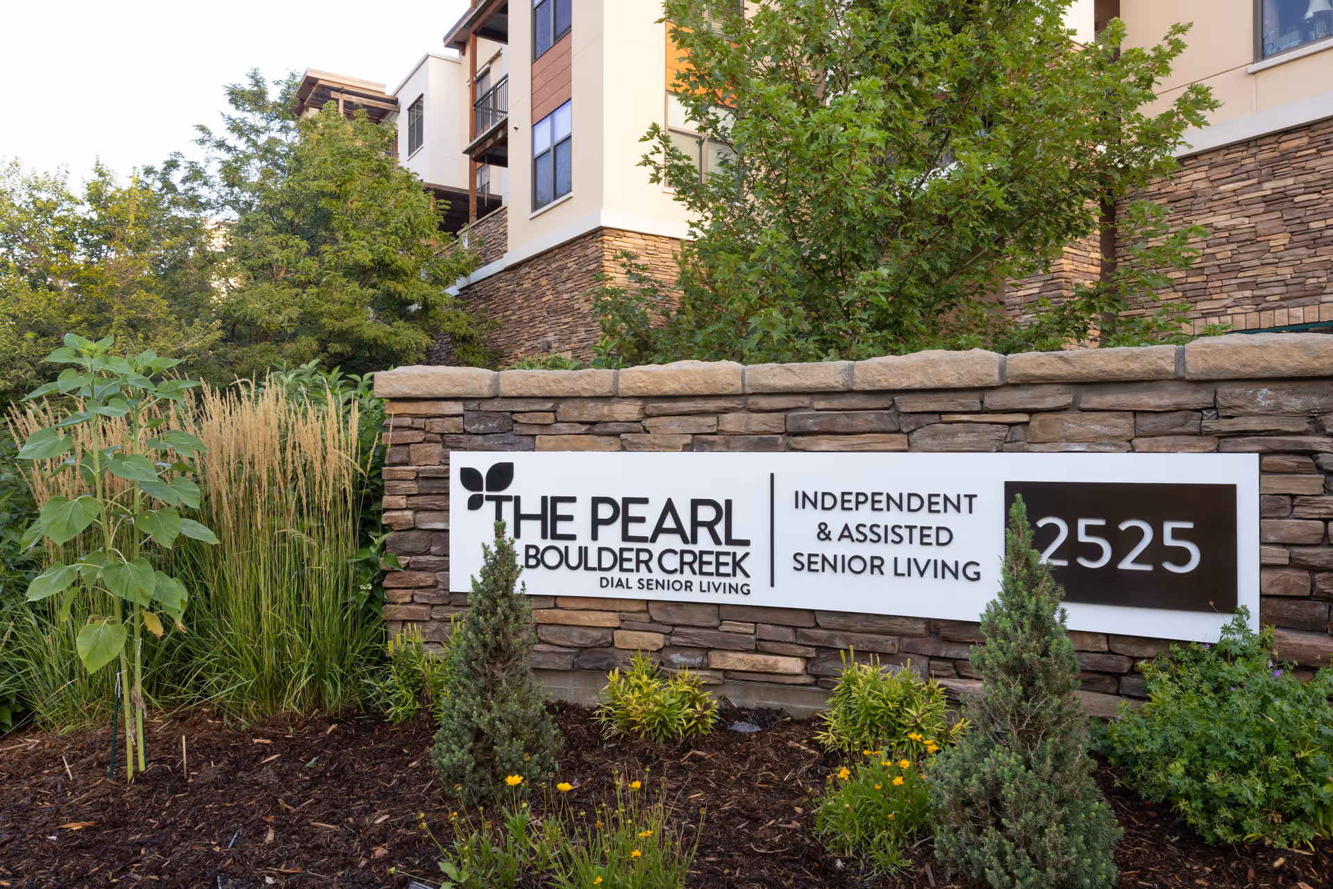 Stone entrance sign for The Pearl Boulder Creek reading 'Independent & Assisted Senior Living 2525' amid landscaping with the building behind.