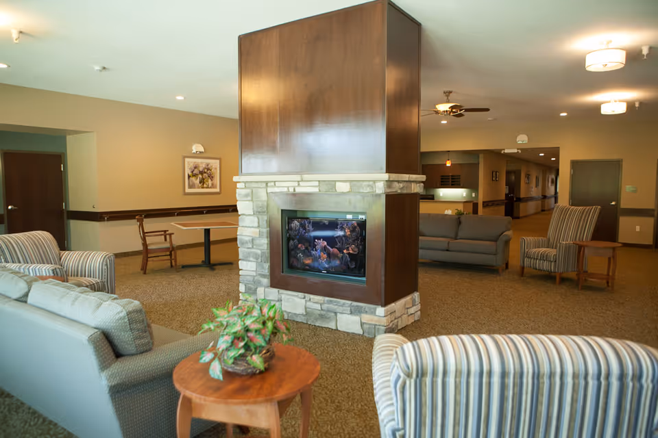 A cozy living room area with a central stone and wood fireplace. The room features multiple seating options including striped armchairs, a gray sofa, and a wooden table with a plant on it. The walls are painted beige with framed artwork, and there are ceiling lights and a ceiling fan. A hallway and additional seating are visible in the background.