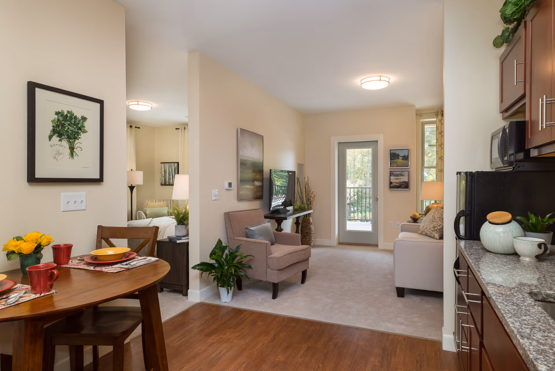 Interior view of a senior living apartment showing a small dining area with a wooden table set for two with red mugs and yellow bowls, a living room with an armchair, sofa, wall-mounted TV, and a door leading to a balcony. The kitchen area with granite countertops and dark wood cabinets is visible on the right. The walls are light beige, and there are framed pictures and plants decorating the space.