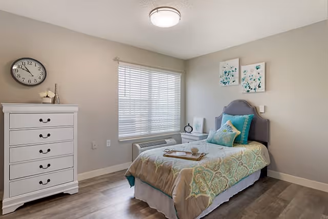 A neatly arranged bedroom with a single bed featuring a gray headboard, teal and beige bedding, and decorative pillows. There is a white nightstand with a clock and a small piece of art on it next to the bed. A white chest of drawers with a clock and decorative items on top is positioned against the wall. A window with white blinds allows natural light into the room, which has light-colored walls and wood flooring.