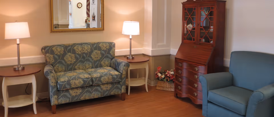 A cozy seating area with a patterned loveseat between two table lamps, a wooden secretary cabinet and an upholstered armchair.