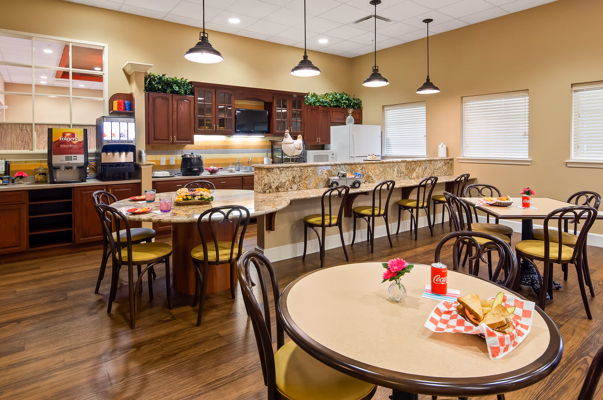 Bright communal dining area with round tables and chairs, a granite serving counter with barstools, and a kitchenette in the background.