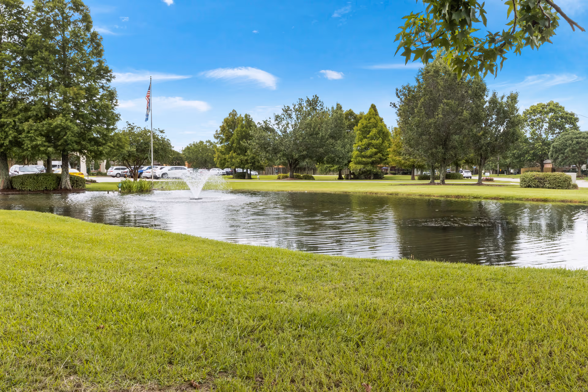 A peaceful outdoor scene at Landmark of Baton Rouge featuring a small pond with a water fountain in the center, surrounded by green grass and trees under a blue sky with scattered clouds. Several parked cars and an American flag on a flagpole are visible in the background.