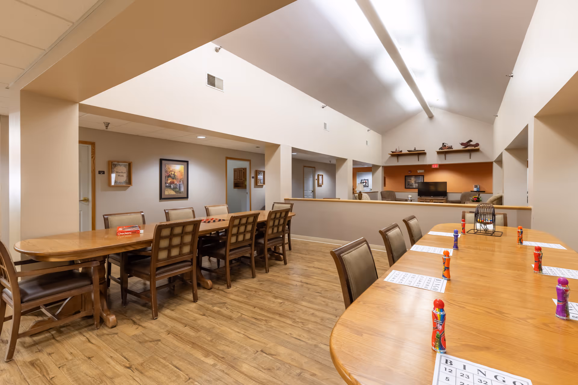 A spacious common area in a senior living facility with two long wooden tables and chairs arranged for group activities. One table has bingo cards and colorful bingo daubers set up, while the other table has a checkers board and pieces. The room has wood flooring, beige walls, and a high ceiling with bright lighting. In the background, there is a seating area with a TV and decorative shelves on the wall.