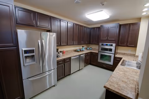 A modern kitchen with dark wood cabinets, a stainless steel refrigerator with a water and ice dispenser, a built-in oven, a dishwasher, a double sink, and beige countertops. The kitchen is well-lit with ceiling lights and has a clean, organized appearance.