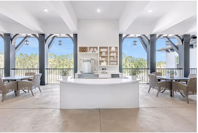Covered outdoor seating area with a central white service counter, wicker tables, and arched openings overlooking trees.