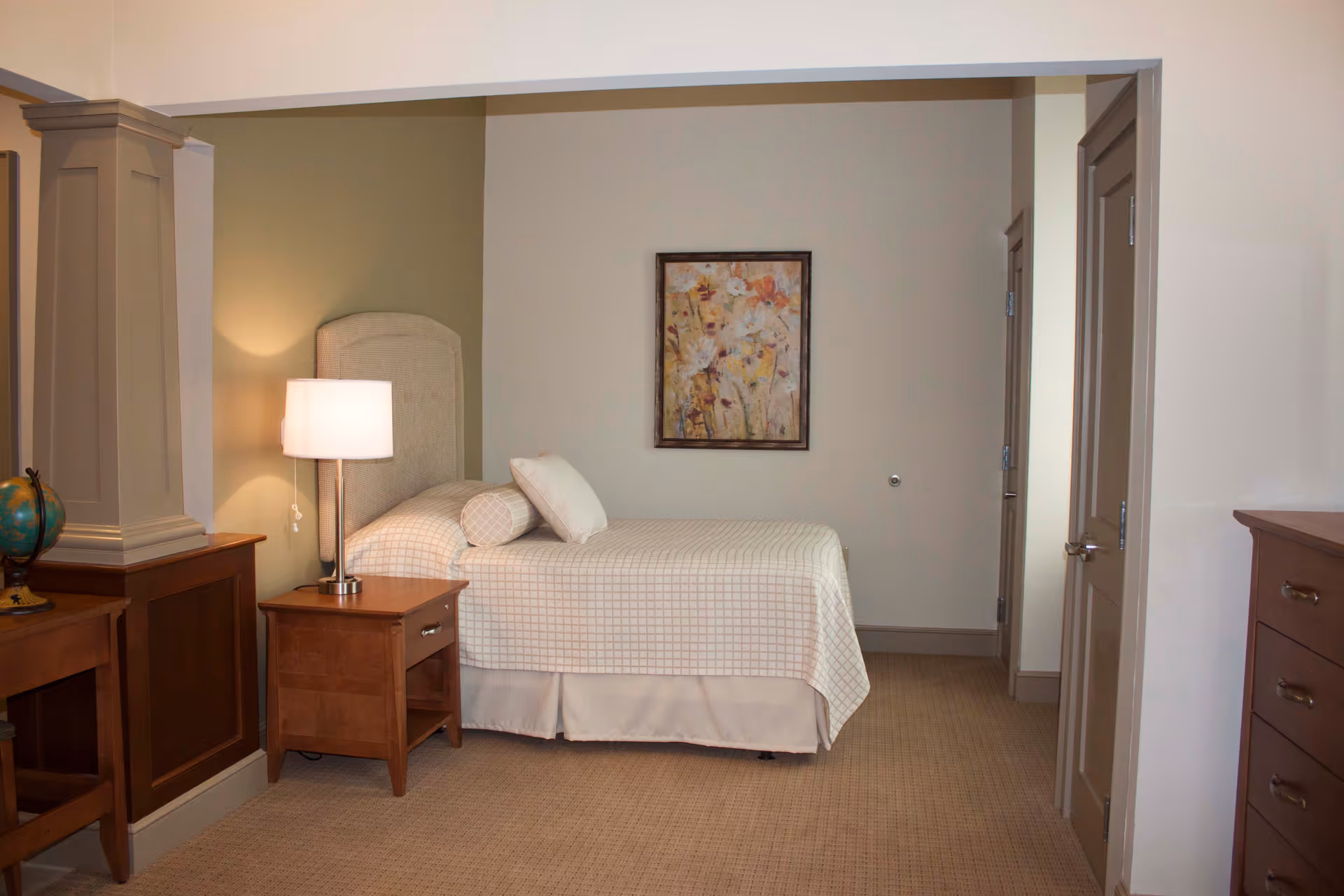 A neatly made single bed with a checkered bedspread and two pillows in a senior living bedroom. Next to the bed is a wooden nightstand with a lit table lamp. A framed floral painting hangs on the wall above the bed. The room has beige carpeting and neutral-colored walls, with a wooden dresser and a small desk with a globe on it visible.