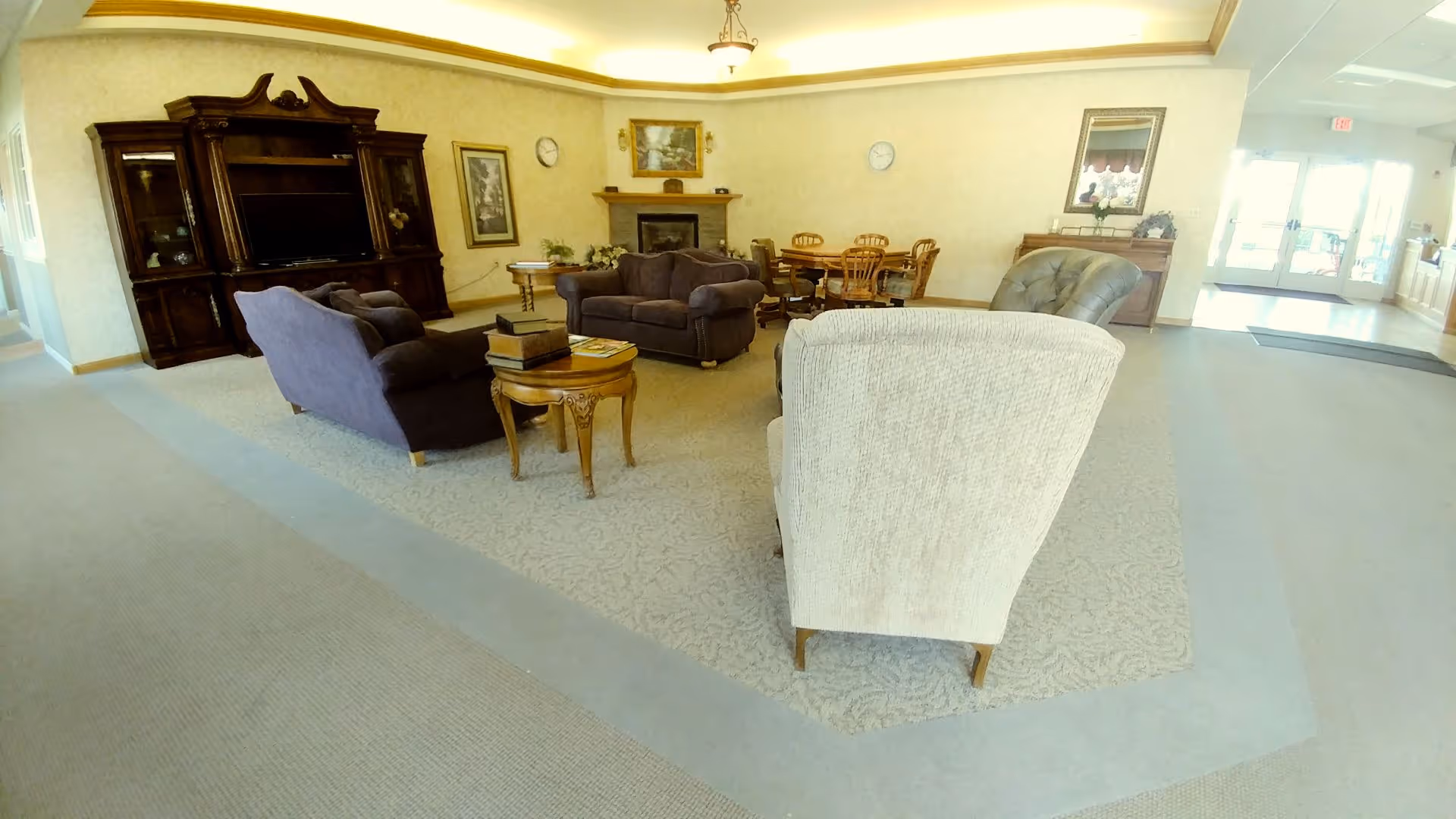 A spacious living room area in a senior living facility featuring a beige armchair in the foreground, two dark brown sofas, a wooden coffee table with books, a large wooden entertainment center with a TV, a fireplace with a painting above it, a round dining table with four chairs, and a sideboard with a mirror and flowers. The room has light-colored walls and carpet, with ceiling lights and an exit door visible in the background.