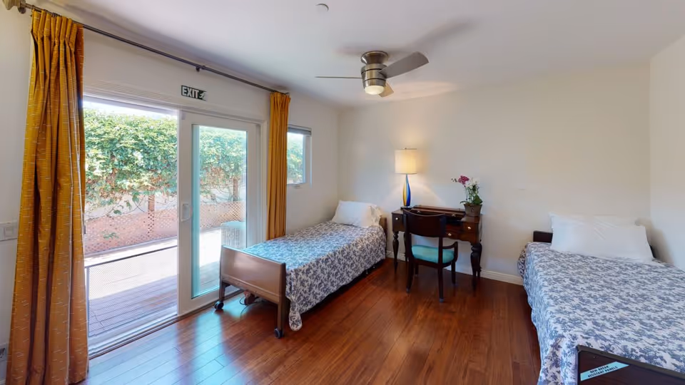 A bright bedroom with two single beds covered in blue and white patterned bedspreads. Between the beds is a wooden desk with a chair, a table lamp, and a small potted plant. The room has wooden flooring, white walls, a ceiling fan, and a large sliding glass door with yellow curtains leading to an outdoor area with greenery.