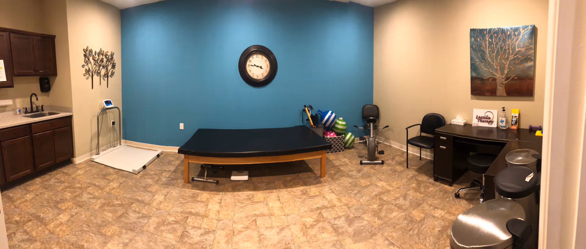 A therapy or medical examination room with a blue accent wall featuring a clock. The room contains a black examination table, an exercise bike, a desk with chairs, and various therapy equipment including exercise balls and weights. There is a sink with cabinets on the left side and a decorative tree wall art on the right wall.