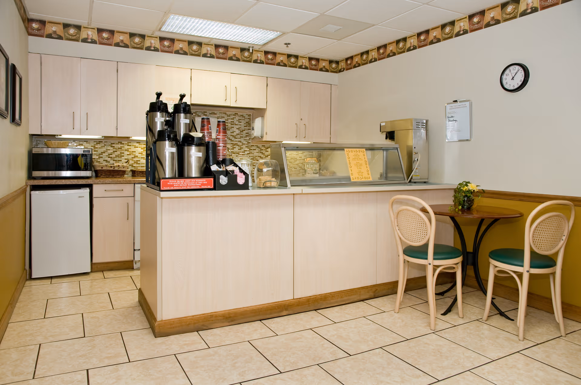 Small kitchenette area with light wood cabinets, a countertop with coffee dispensers, a microwave, a small refrigerator, and a display case. There is a small round table with two chairs with green cushions and a small plant on the table. A clock and a whiteboard are mounted on the wall.