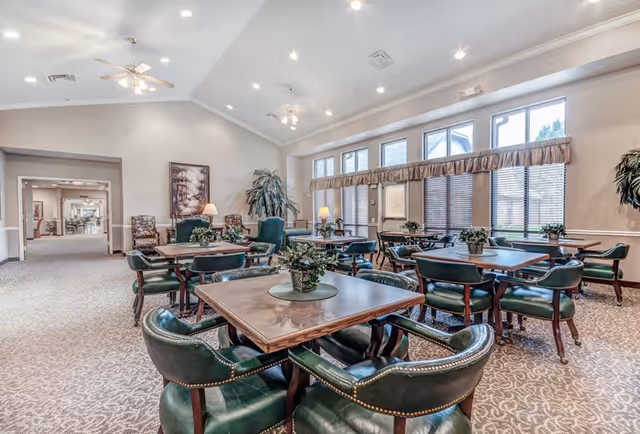 A spacious, well-lit common area with multiple wooden tables surrounded by green leather chairs. The room features large windows with blinds and valances, ceiling fans, recessed lighting, carpeted floors, and decorative plants on the tables and in the corners. There is a hallway visible in the background leading to other parts of the facility.