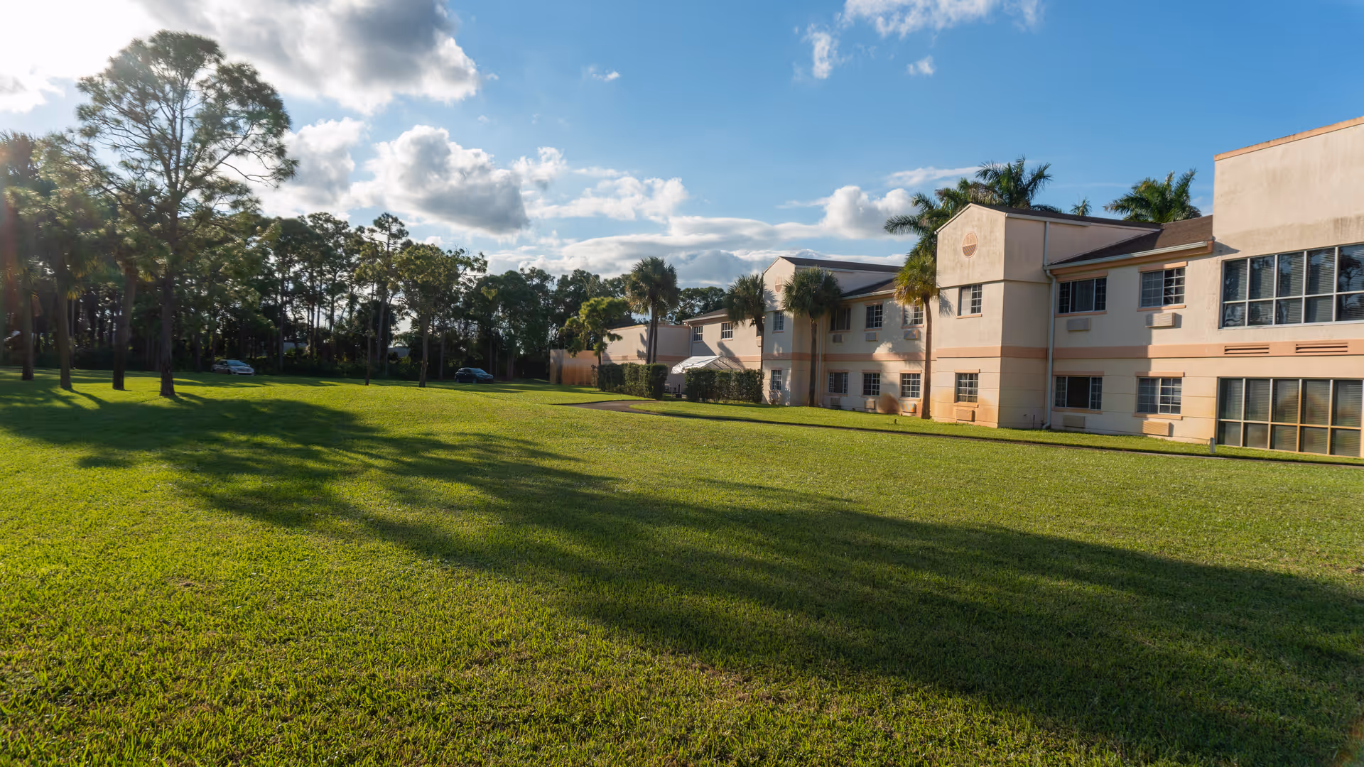 A wide green lawn with trees and palm trees in front of a two-story beige senior living building under a partly cloudy sky.