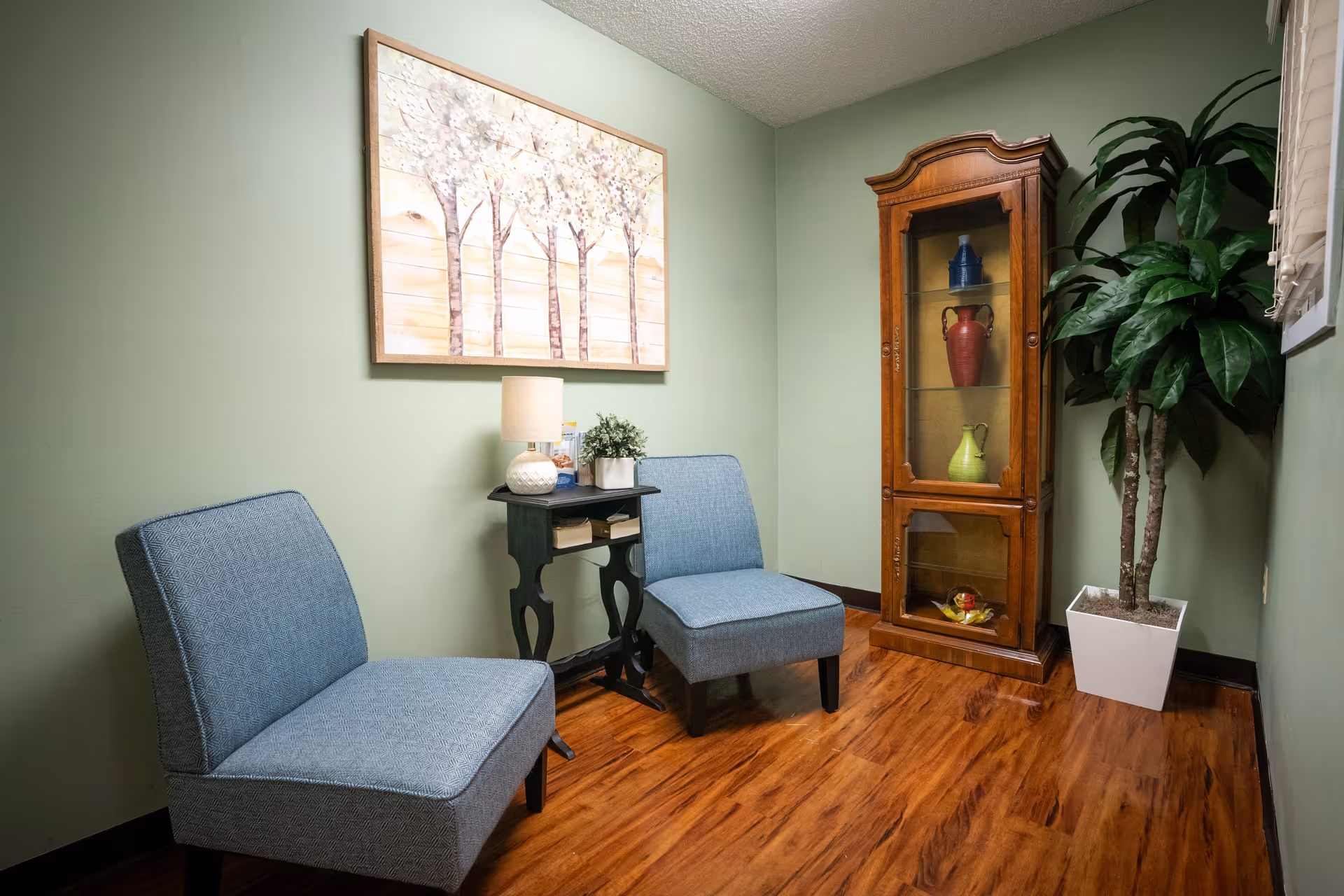 A small sitting area with two blue upholstered chairs, a black side table with a lamp and a small plant, a wooden display cabinet with decorative vases, a large potted plant, and a painting of trees on a light green wall.