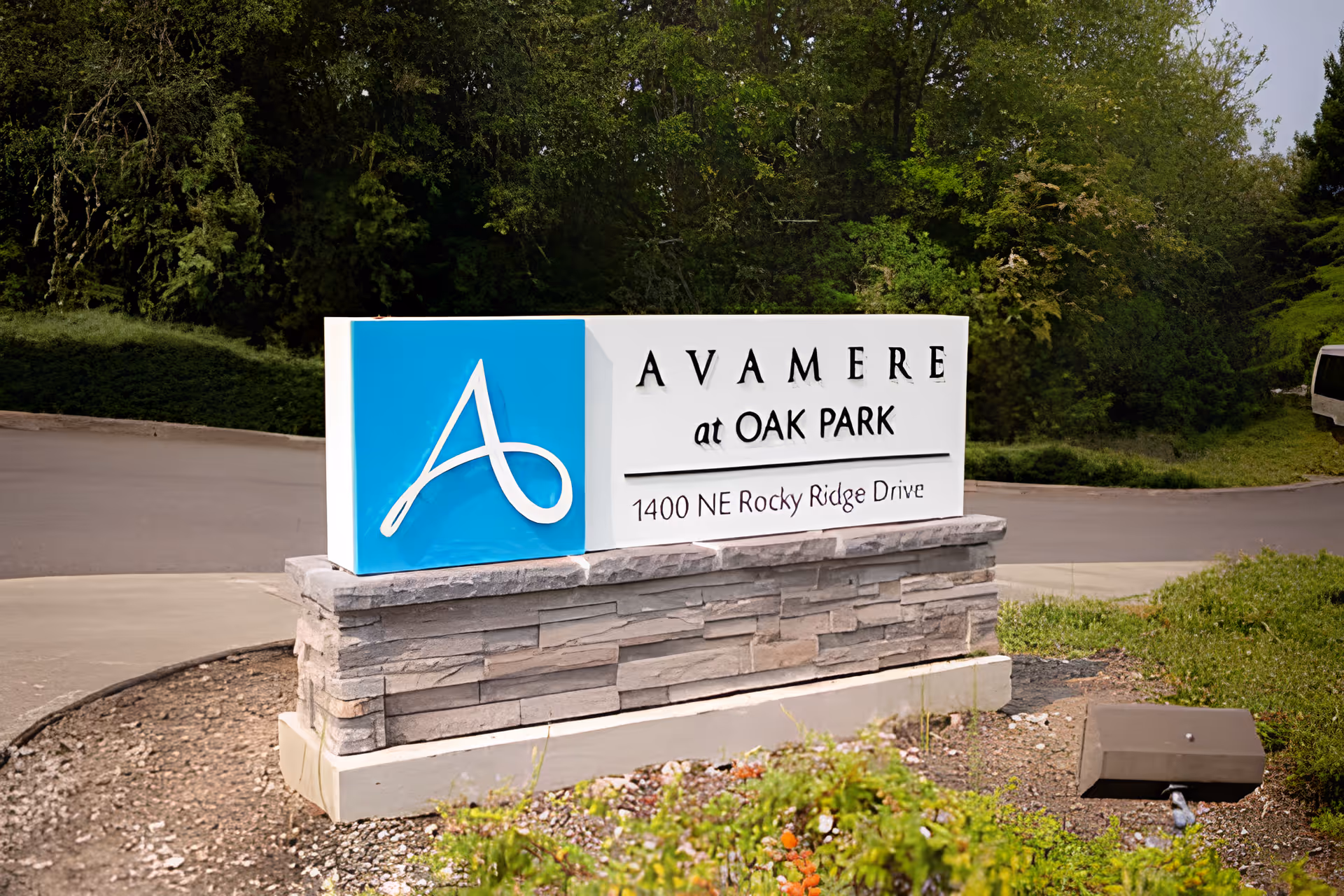 A stone and concrete sign for Avamere at Oak Park located at 1400 NE Rocky Ridge Drive, surrounded by greenery and trees with a road in the background.