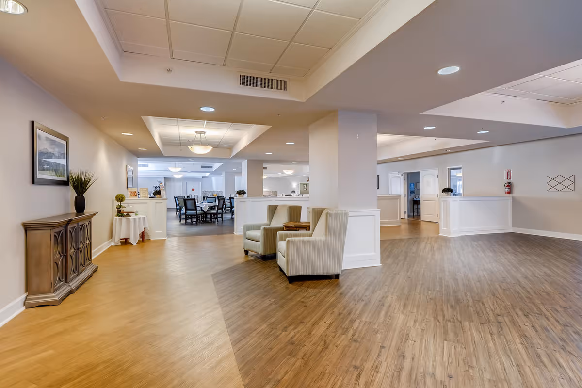 Spacious interior common area with wood flooring, two beige armchairs with a small table between them, a wooden sideboard with a plant on top, and a dining area with tables and chairs in the background. The ceiling has recessed lighting and a central light fixture.