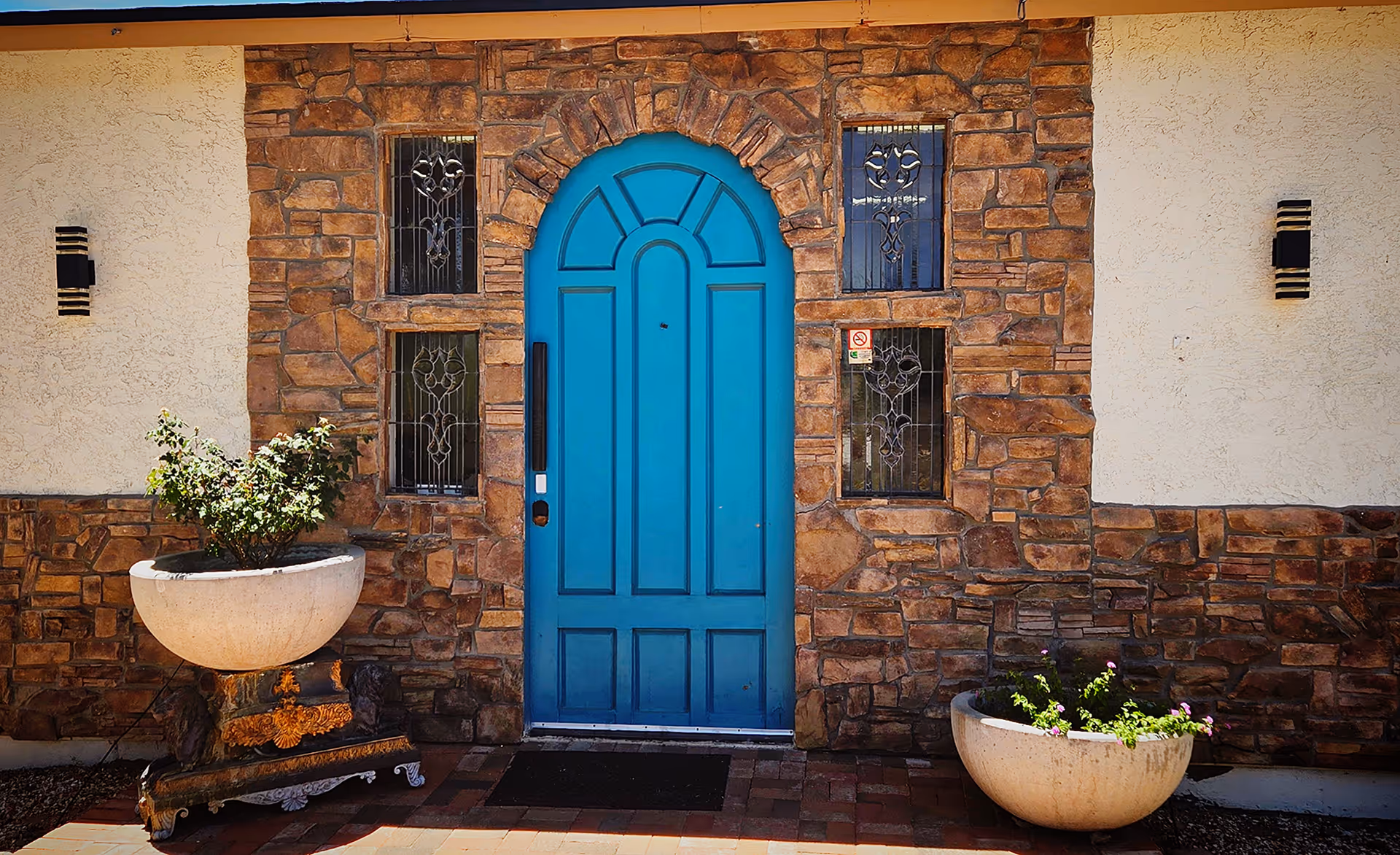 Front entrance of a building with a bright blue arched door set in a stone wall. There are four decorative glass windows around the door and two large round planters with green plants on either side of the entrance. The wall is a combination of stone and stucco with two modern black wall lights.